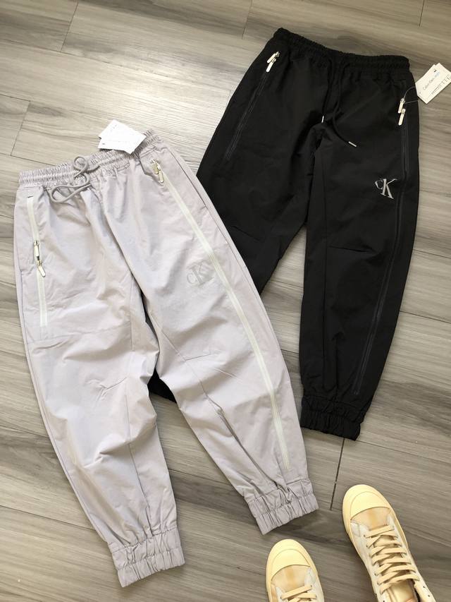 CK Jeans SS24 滑板系列 字母胶印 长拉链重工男女款运动休闲束脚长裤. 颜色：灰色 黑色. 面料：定织涂层科技面料. 工艺：印花 立体剪裁. 尺码：